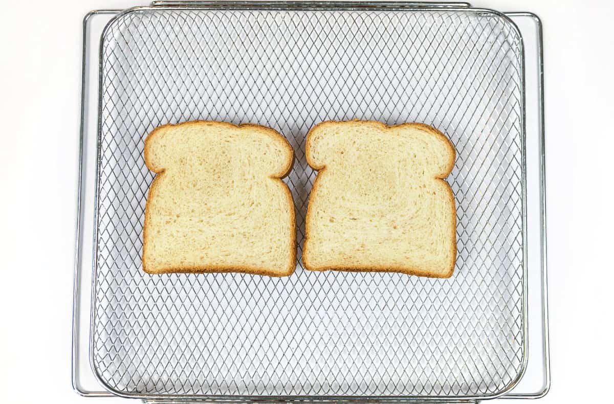 Two slices of bread on the air fryer basket before toasting.