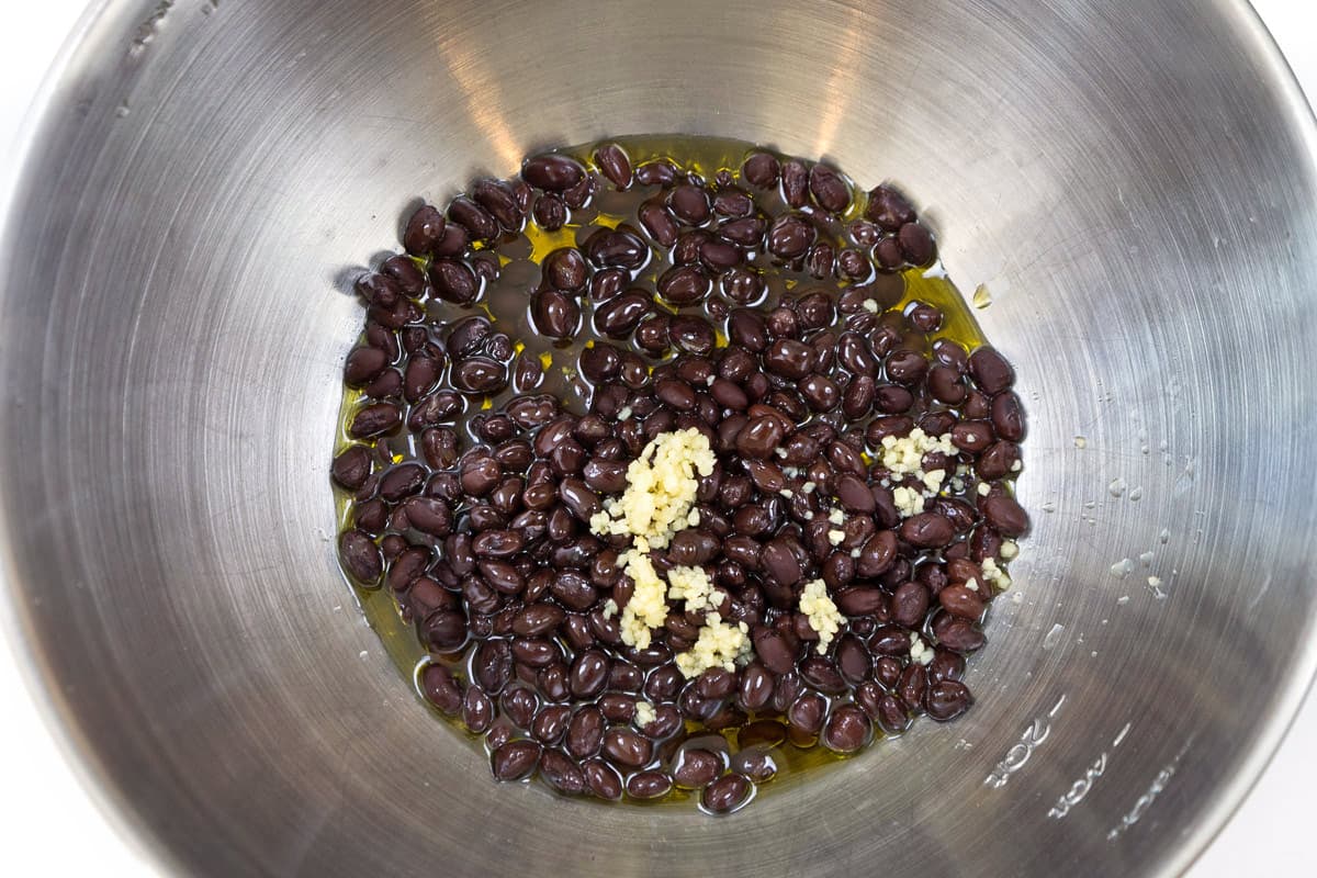 Olive oil and minced garlic are added to the black beans in a bowl.