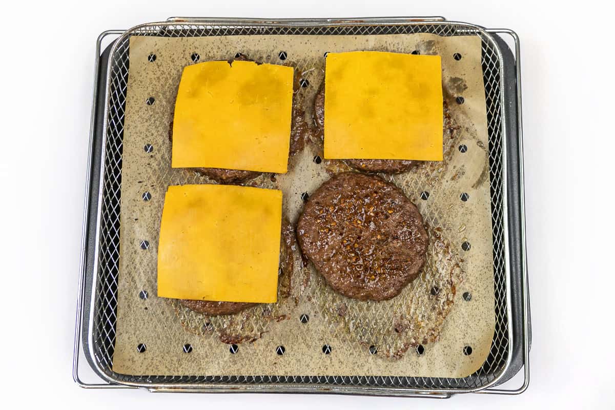 Four cooked hamburger patties with melted cheese on parchment paper on the air fryer basket.