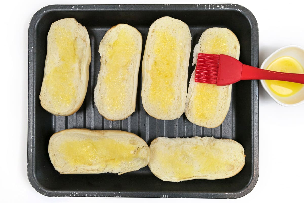 Buttered hoagie rolls on a broiler pan.